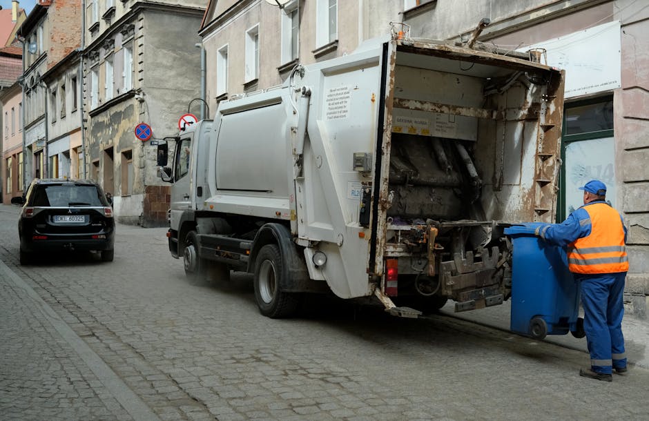 A large urban outdoor scene showing an overflowing rubbish collection point with multiple black and red waste bins, a grey mixed paper and card recycling container, and various discarded items spillin