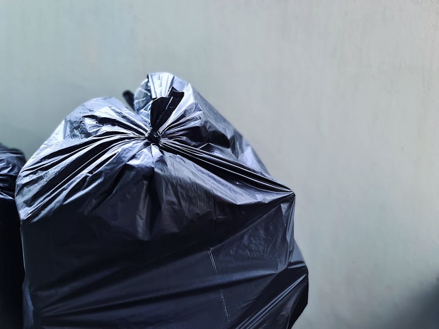 Close-up image of a black plastic rubbish bag, tightly sealed at the top with a twist tie, placed on a flat surface against a plain, light-colored wall. The bag appears full, with visible creases and