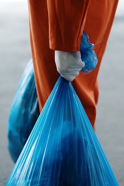 Close-up image of a black plastic rubbish bag, tightly sealed at the top with a twist tie, placed on a flat surface against a plain, light-colored wall. The bag appears full, with visible creases and